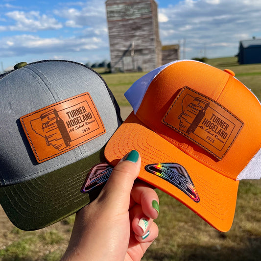 Two hats, one gray and one orange, held by a hand with a blurred outdoor background.